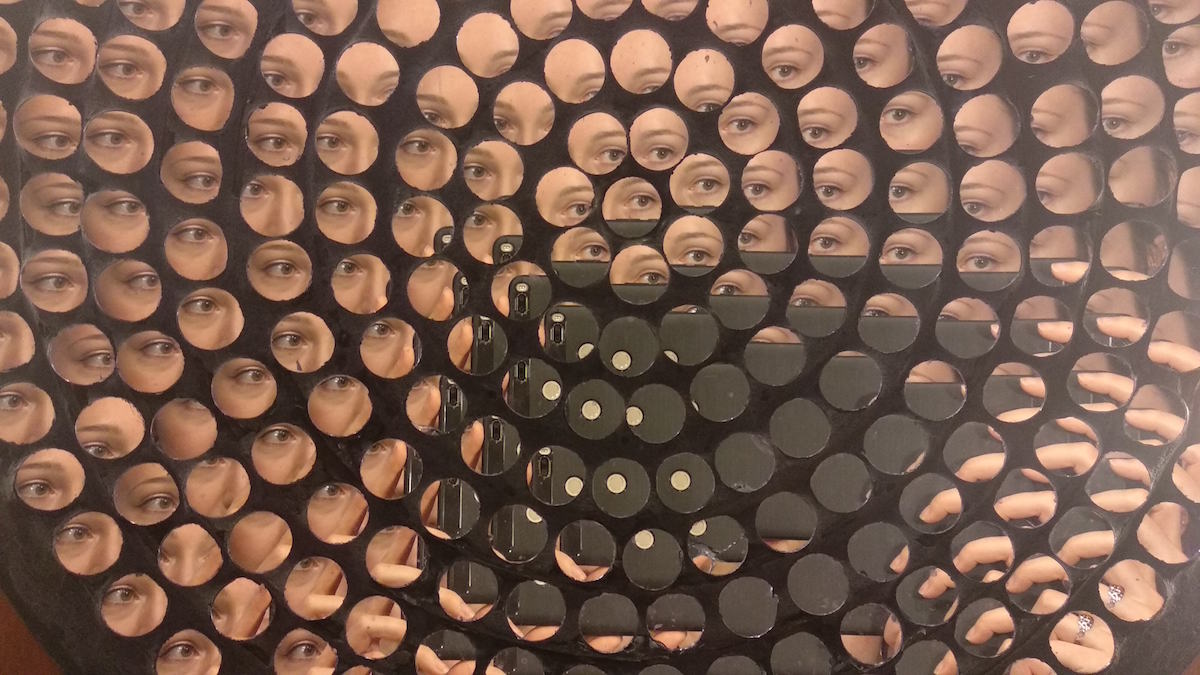 Image description: Concentric circles of small round mirrors set in a wooden board, showing a kaleidoscopic swirl of slightly-skewed views of someone's eye and the cell phone they are holding up as though to take a mirror selfie.